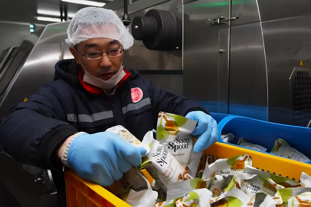 A worker in protective gear sorts soup packages in a factory. Wearing gloves, a hairnet, and a mask, he exhibits focus and careful handling.