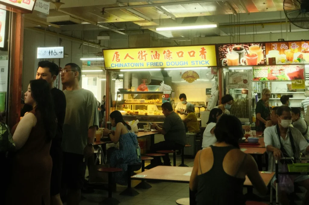 A busy food court scene with people seated and standing near a stall with a yellow sign reading "Chinatown Fried Dough Stick." The atmosphere is lively.