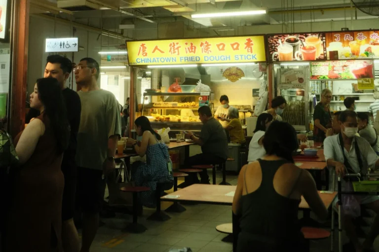 People sit and stand in a bustling food court with a bright sign reading "Chinatown Fried Dough Stick." Warm lighting and a lively atmosphere.