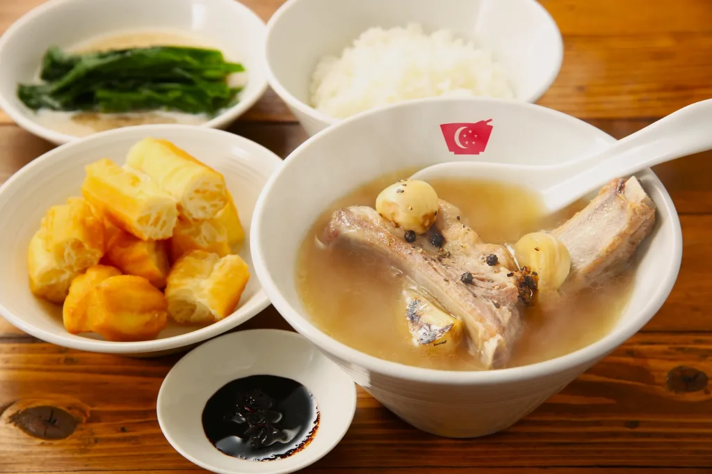 A bowl of Bak Kut Teh with pork ribs, black pepper, and garlic in broth, served with white rice, fried dough fritters, and soy sauce on a wooden table.