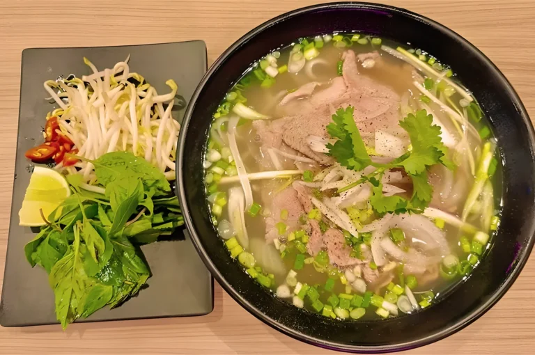 A steaming bowl of pho with beef, fresh greens, and bean sprouts, garnished with cilantro. A side plate holds lime wedges, basil, and chili slices.