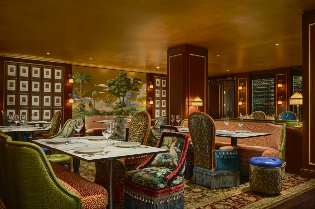 Elegant restaurant interior with mural of trees and a building. Plush upholstered chairs, white tablecloths, soft lighting, and cozy ambiance.