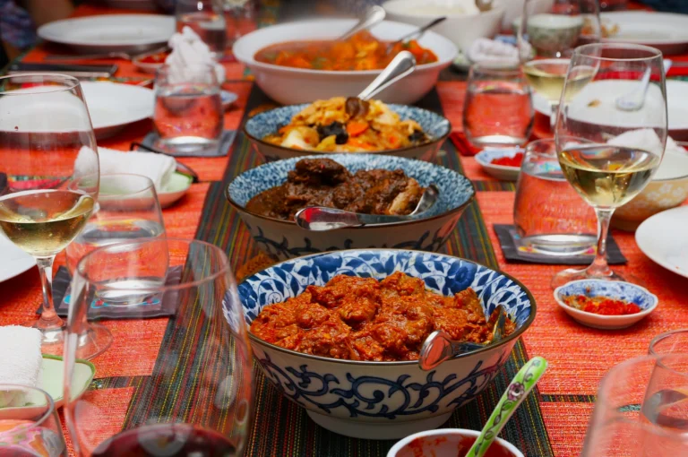 A colorful dining table with blue-patterned bowls of curry dishes, surrounded by wine glasses and plates. The setting is inviting and festive.