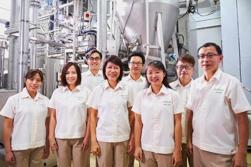 A group of eight people in matching white shirts and beige pants smile inside an industrial factory with metal machinery in the background.