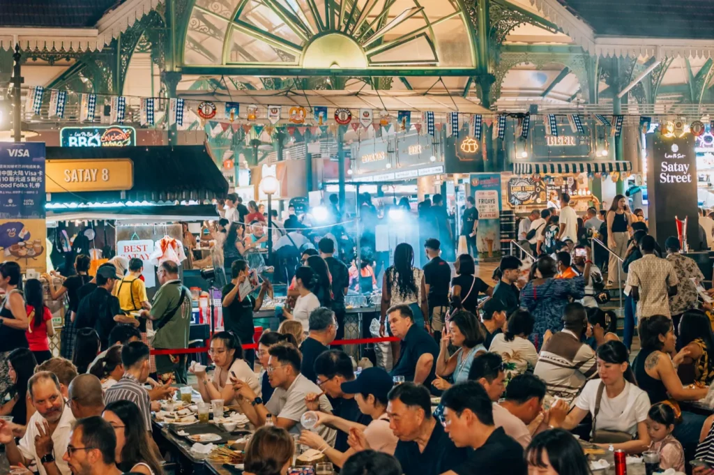 Crowded vibrant hawker center, filled with people at tables enjoying food. Colorful stalls and smoke create a lively and bustling atmosphere.