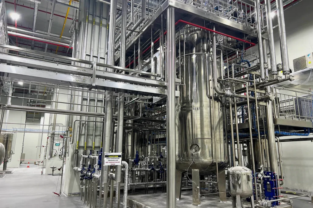 Industrial interior with large stainless steel tanks and extensive piping network. The space is clean, orderly, and brightly lit, conveying precision.