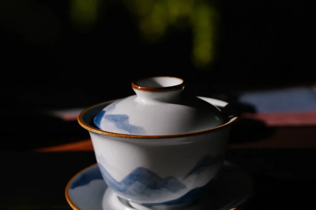 A porcelain teacup with a lid features delicate blue mountain designs and gold rims, set against a dark, blurred background. It evokes a serene, elegant atmosphere.