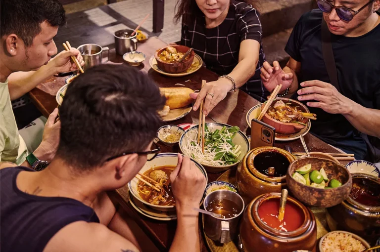 Four people sit around a wooden table enjoying Asian cuisine. There are bowls of noodles, herbs, and colorful sauces, creating a vibrant, communal atmosphere.
