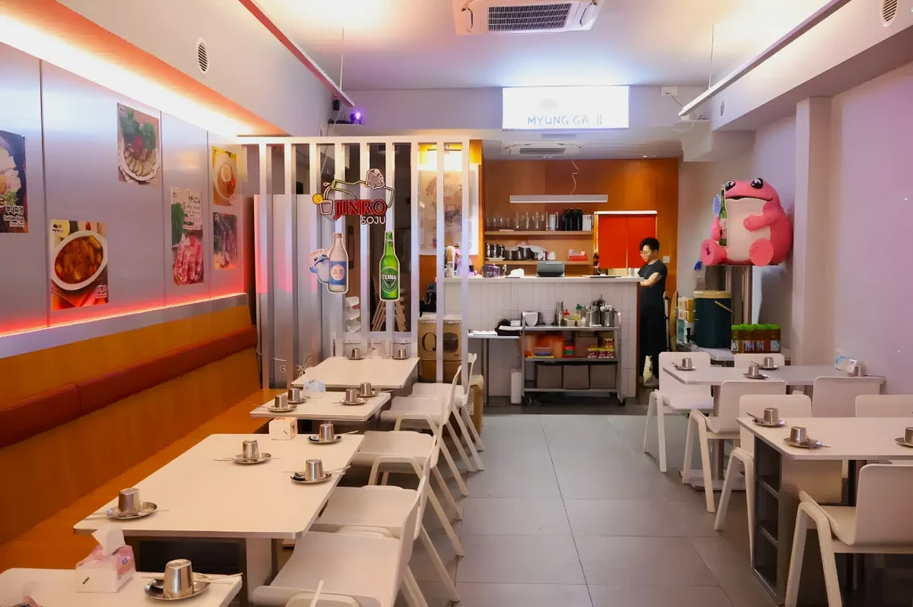 Cozy restaurant interior with white tables and chairs, metal hot pot sets, and a row of booths on the left. A staff member stands near the counter, and a large pink plush character adds whimsy to the corner. Soft, warm lighting enhances the inviting atmosphere.