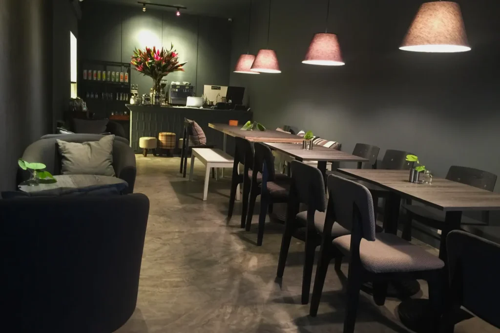 Cozy café interior with dim lighting, featuring gray walls and floors. Wooden tables with black chairs line the right, and cushioned seating is on the left. A counter at the back has a flower arrangement.