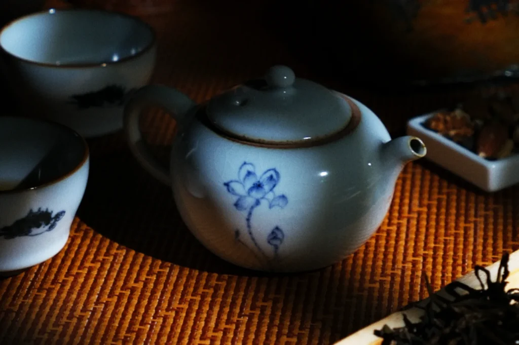 Small white teapot with a blue floral design on a woven mat, surrounded by tea cups and a dish. Dim, cozy lighting enhances a calm atmosphere.