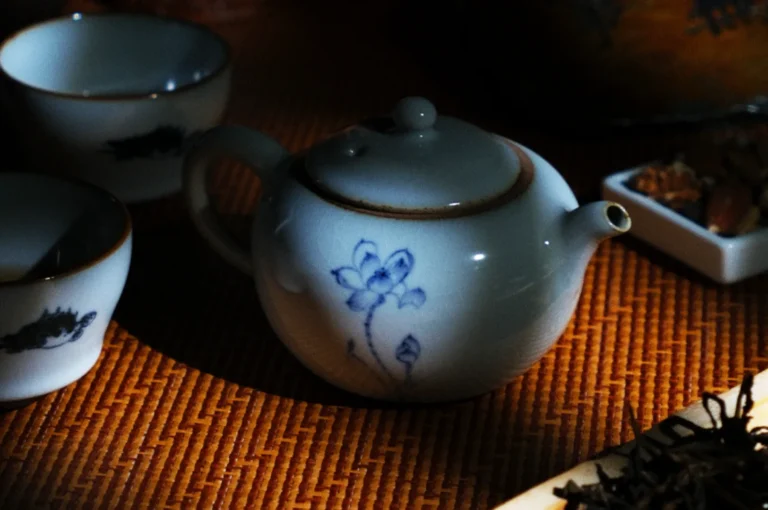 Small white teapot with a blue floral design on a woven mat, surrounded by tea cups and a dish. Dim, cozy lighting enhances a calm atmosphere.