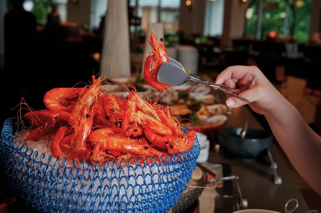 A hand is using tongs to pick a bright orange shrimp from a basket filled with ice and seafood. The setting is a vibrant buffet with various dishes.
