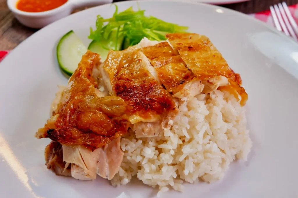 Plate of Hainanese chicken rice with sliced, golden-brown chicken on white rice. Garnished with cucumber and celery, with a small bowl of chili sauce.