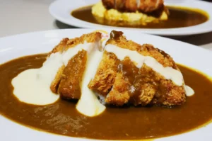 Crispy breaded cutlet drizzled with melted cheese sauce atop fluffy rice, surrounded by rich brown curry on a white plate; comfort and warmth.