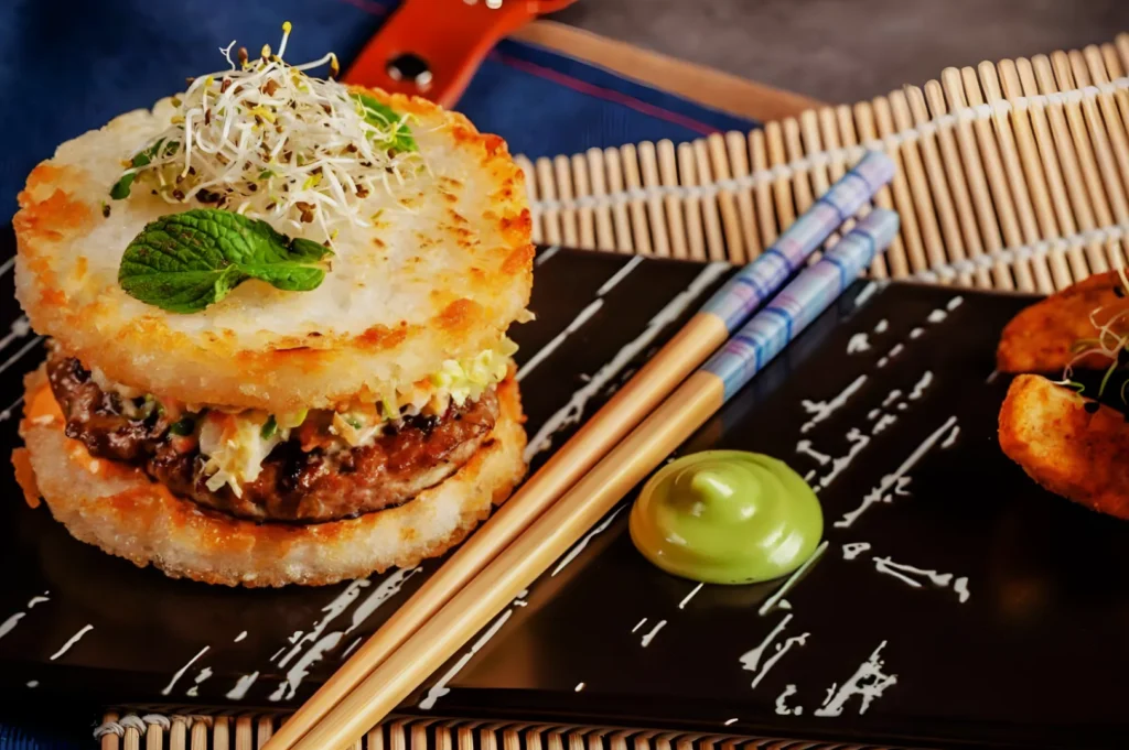 A rice burger filled with beef and slaw is topped with sprouts and mint on a black plate. Nearby are chopsticks and a dollop of green sauce.