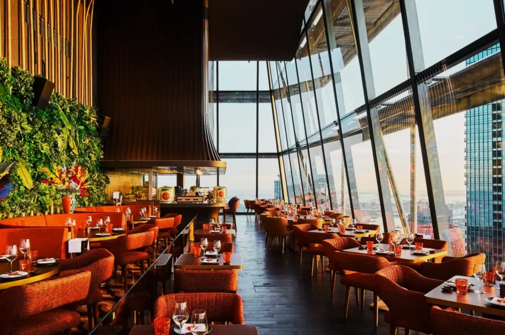 Modern restaurant interior with tall glass walls offering a city view. Warm lighting, orange chairs, wooden tables set with glasses, cutlery, lush green plant wall.