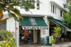 Street view of a cozy bakery with green awnings labeled "Tiong Bahru Bakery." There are plants outside, and two people are seated inside, creating a relaxed atmosphere.