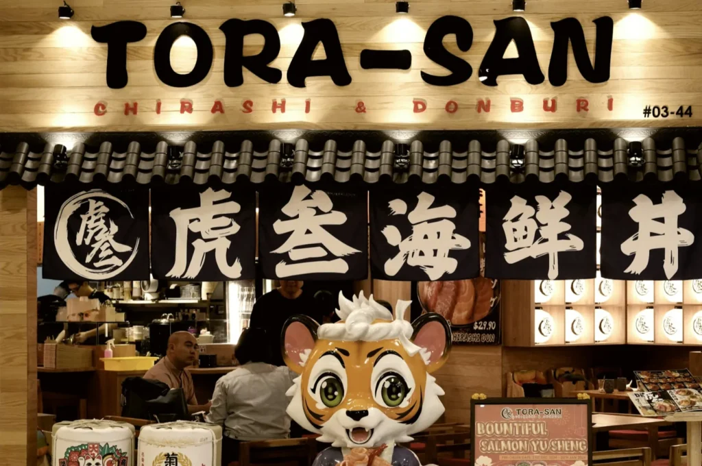 Restaurant entrance of Tora-San, featuring a large cartoon tiger statue. Traditional Japanese banners, warm lighting, and patrons inside.