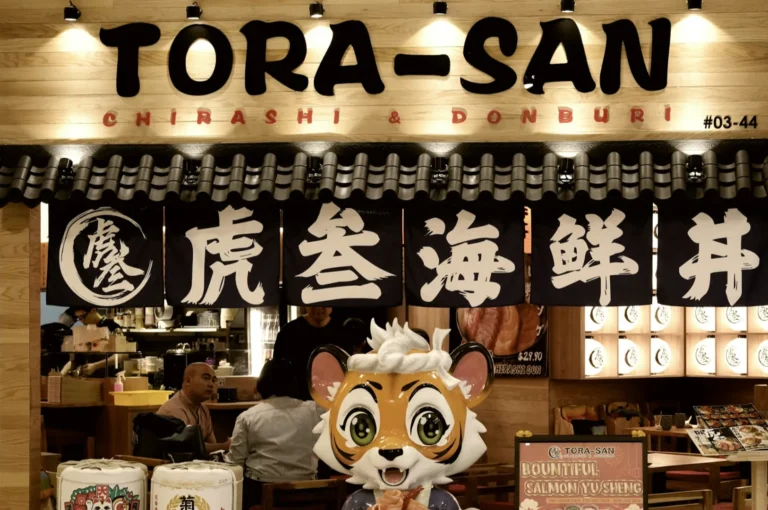 Restaurant entrance of Tora-San, featuring a large cartoon tiger statue. Traditional Japanese banners, warm lighting, and patrons inside.