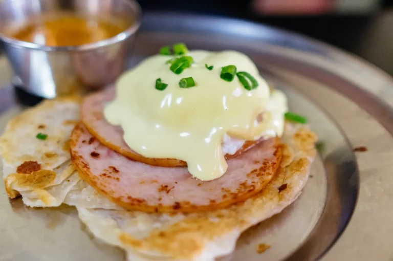 Eggs Benedict on crispy flatbread, topped with ham, creamy hollandaise sauce, and chopped green onions, with sauce in a metal cup nearby.