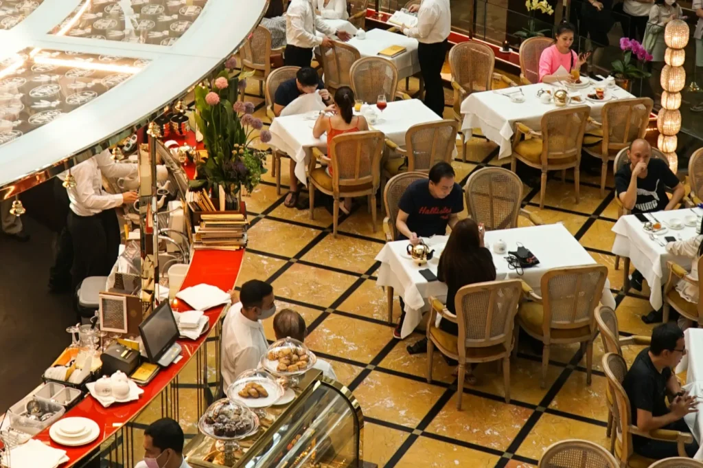 A bustling cafe with patrons seated at tables with white tablecloths, enjoying meals. Waitstaff in white uniforms serve guests. Bright, lively atmosphere.