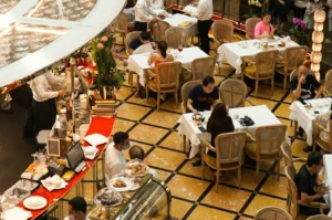 A bustling cafe with patrons seated at tables with white tablecloths, enjoying meals. Waitstaff in white uniforms serve guests. Bright, lively atmosphere.