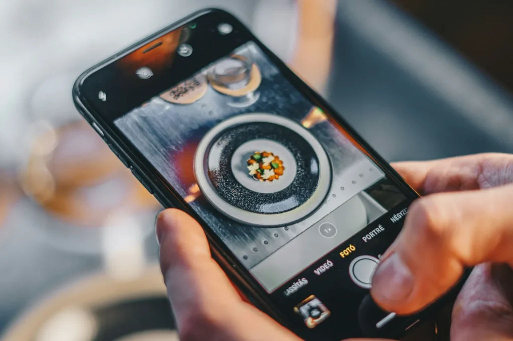A person photographs a gourmet dish with their smartphone. The dish appears colorful and artfully arranged, emphasizing a modern culinary experience.