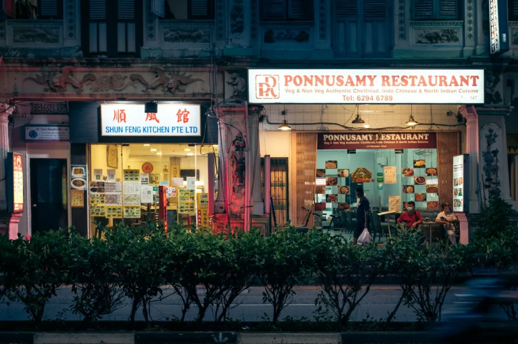 Nighttime street view of two side-by-side lit restaurants in Singapore. "Shun Feng Kitchen" on the left and "Ponnusamy Restaurant" on the right. Lush greenery in the foreground.