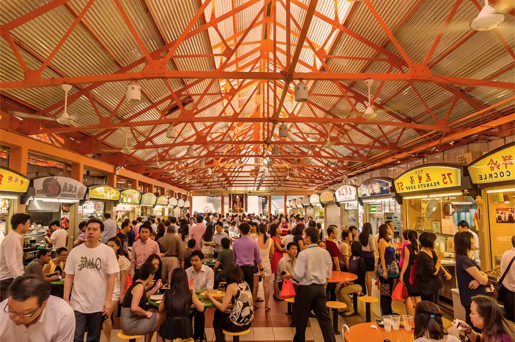 Crowded indoor food market with bustling patrons. Bright, lively atmosphere with colorful signage and high, exposed steel ceiling beams.