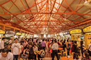 Crowded indoor food market with bustling patrons. Bright, lively atmosphere with colorful signage and high, exposed steel ceiling beams.
