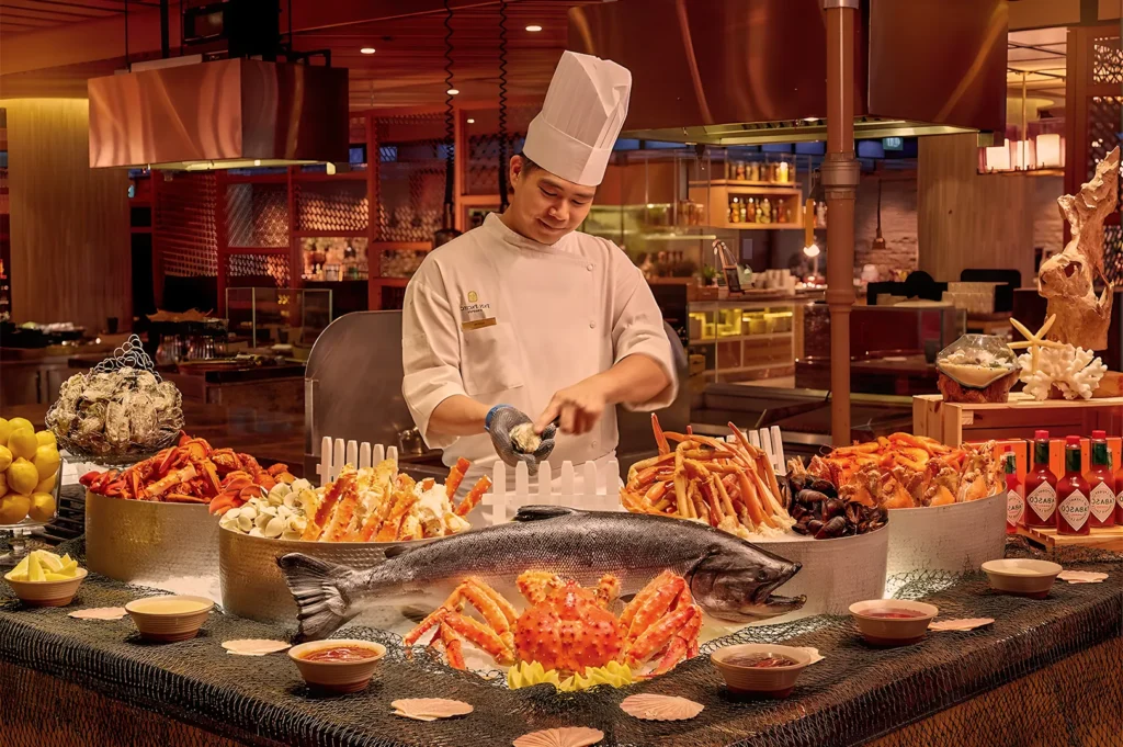 A chef in a white uniform and hat prepares seafood at a lavish buffet. The table displays a large fish, crab, mussels, and condiments, evoking luxury.