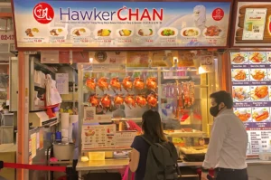 A food stall named Hawker Chan displays roast meats in a glass case. Two customers stand in front, one with a backpack. The atmosphere is busy and vibrant.