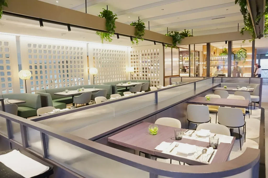 Modern restaurant interior with soft lighting, green and gray seating, and tables set with white linens. Hanging plants add a fresh touch. Calm, inviting atmosphere.