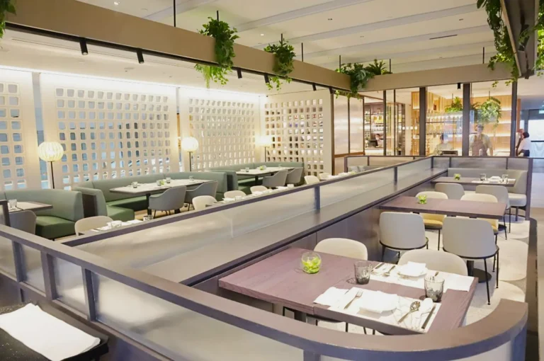 Modern restaurant interior with soft lighting, green and gray seating, and tables set with white linens. Hanging plants add a fresh touch. Calm, inviting atmosphere.