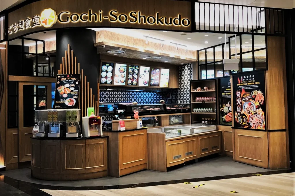 A Japanese restaurant called Gochi-So Shokudo features a colorful and inviting storefront with illuminated menus and food displays, evoking a welcoming atmosphere.