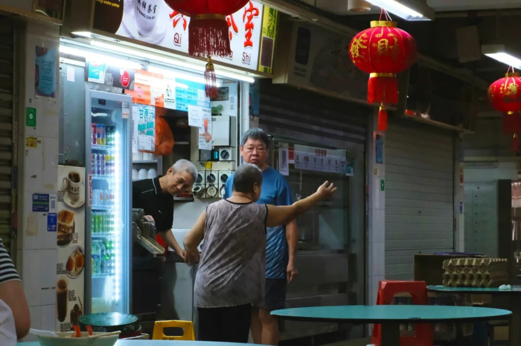 A woman gestures while talking to two men at a dimly lit street food stall adorned with red lanterns. The atmosphere is lively and informal.