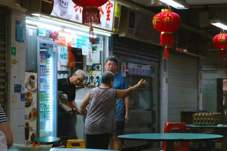 A woman gestures while talking to two men at a dimly lit street food stall adorned with red lanterns. The atmosphere is lively and informal.