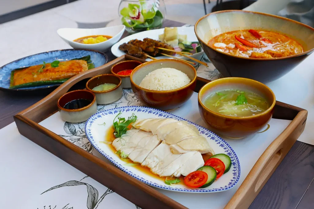 A variety of Asian dishes are arranged on a wooden tray, featuring sliced chicken, rice, broth, and sauces. In the background are satay skewers, a spicy soup, and desserts, creating an appetizing, multicultural spread.