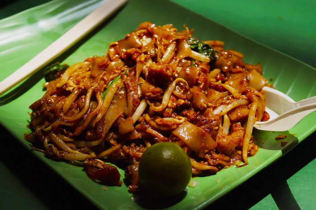 A plate of stir-fried flat noodles with vegetables and meat is served on a green plate. A lime and white utensils are placed beside the dish.