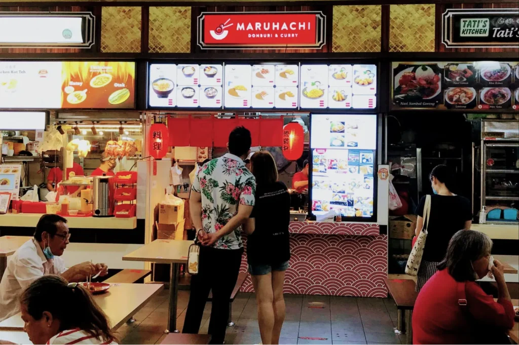 A couple stands together, viewing the menu of "Maruhachi Donburi & Curry." The scene is vibrant with red lanterns. Customers sit at nearby tables eating.