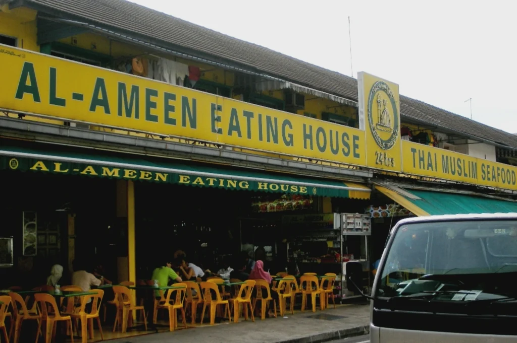 Outdoor view of "Al-Ameen Eating House," a casual Thai Muslim seafood spot. Yellow signage, orange chairs, and people dining create a welcoming vibe.