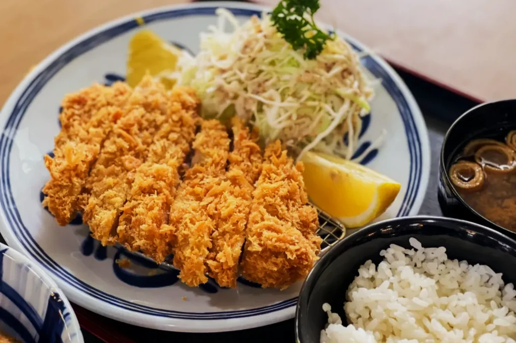 A plate of crispy, golden-brown tonkatsu with shredded cabbage, a lemon wedge, and garnished with parsley. Served alongside bowls of rice and miso soup.