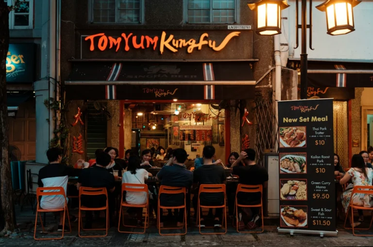 Outdoor dining scene at Tom Yum Kung Fu restaurant, featuring a group of people seated on orange chairs. Warm lights and menu board add a cozy vibe.