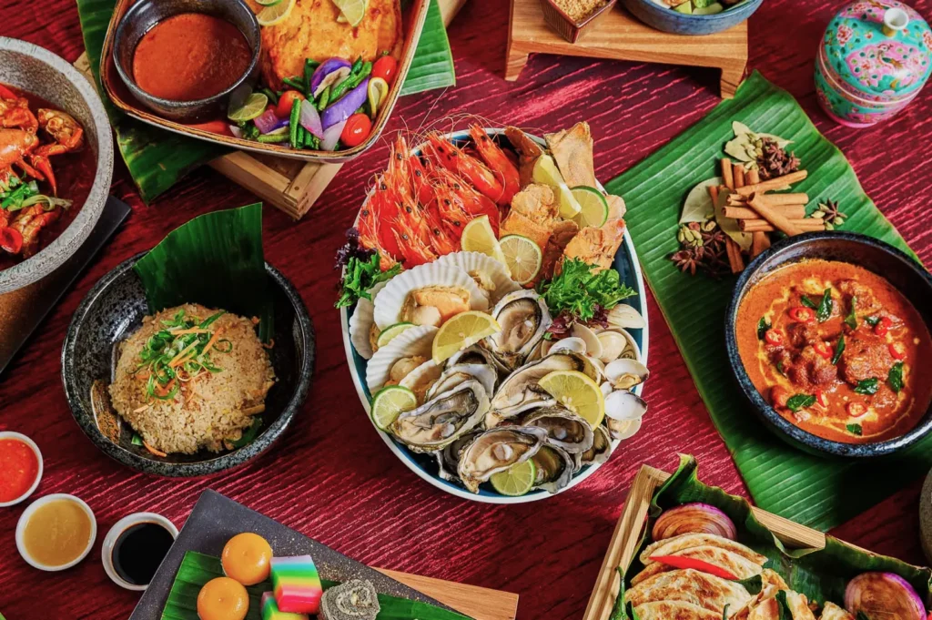 A vibrant spread of Asian cuisine on a red table. Central seafood platter with shrimp, oysters, and lime. Surrounding dishes include curry, rice, sauces, and colorful desserts on banana leaves, creating a festive and appetizing display.