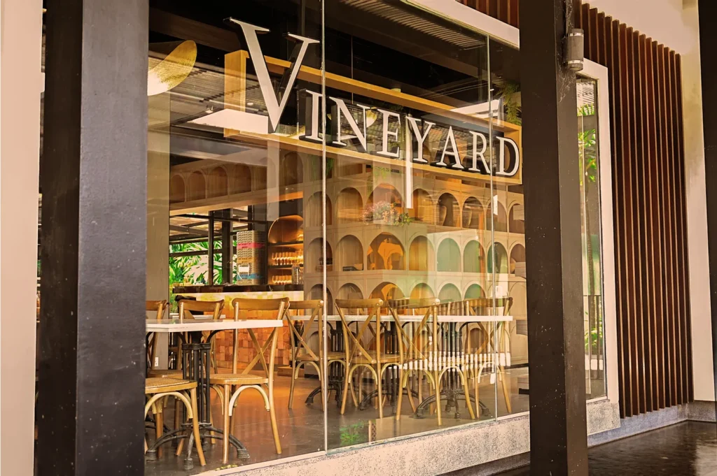 Chic restaurant with "Vineyard" on the window, featuring wooden chairs and tables. Shelves with wine add warmth, creating a cozy and inviting ambiance.