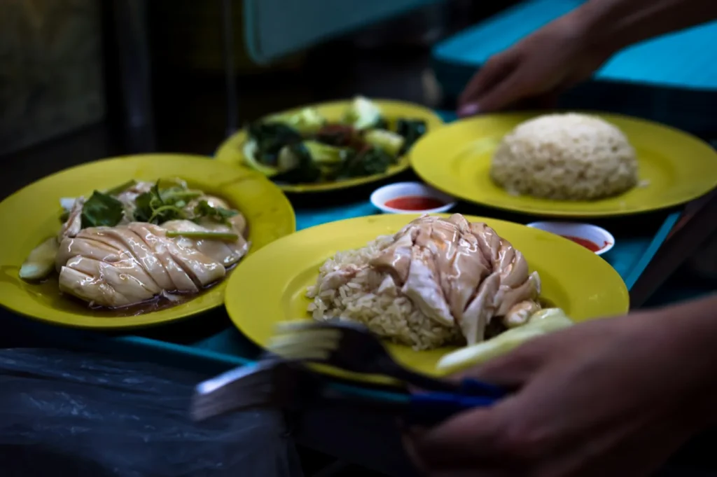 Close-up of yellow plates with Hainanese chicken rice, sliced chicken, and greens. A hand serves food. Small bowls of chili sauce accompany the meal.