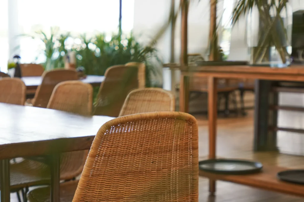 Wicker chairs surround wooden tables in a bright, airy cafe filled with lush green plants, creating a serene and inviting atmosphere.
