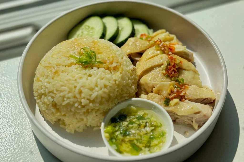 A plate of Hainanese chicken rice with sliced chicken, cucumber slices, and a mound of seasoned rice, garnished with green onions, next to chili sauce.