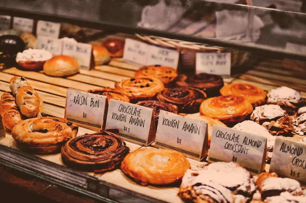 A bakery display with various pastries like raisin buns, chocolate kouign amann, and almond croissants. Warm lighting enhances the inviting, cozy feel.